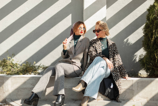 В городе с солнечным светом сидят две красивые стильные подружки модели в модной одежде в леопардовых пальто, клубных джинсах, костюме и туфлях. Городской винтажный стиль и модный наряд - Фото, изображение
