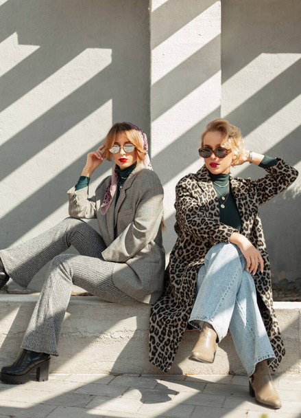 Stylish beautiful young girlfriends in fashionable clothes with leopard coat. jeans, suit, sunglasses and shoes sitting near a concrete wall with sunlight in sunny day. Two urban fashion women - Fotoğraf, Görsel