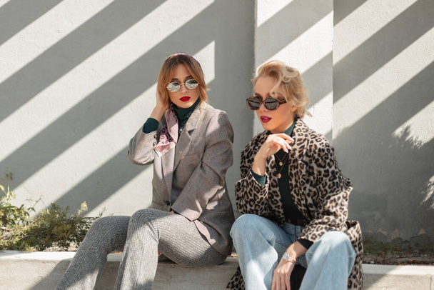 Две красивые гламурные молодые девушки женщины в ретро солнцезащитных очках в стильном наряде в леопардовом пиджаке, костюме, синих джинсах сидят на улице в солнечной линии. Крутые винтажные красивые девушки на открытом воздухе - Фото, изображение