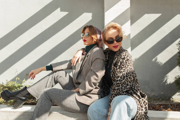 Kaksi nuorta kaunista blondia hipsteriä trendikkäissä syksyn-kevään vaatteissa. Seksikkäät huolettomat naiset istuvat lähellä harmaata betoniseinää auringonvalossa. Naismallit hauskaa - Valokuva, kuva