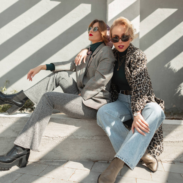 Zwei junge Glamour-Freundinnen in modischen Klamotten sitzen im Sonnenlicht auf der Straße. Mode-Spaß Freundinnen mit Vintage-Sonnenbrille im Freien - Foto, Bild