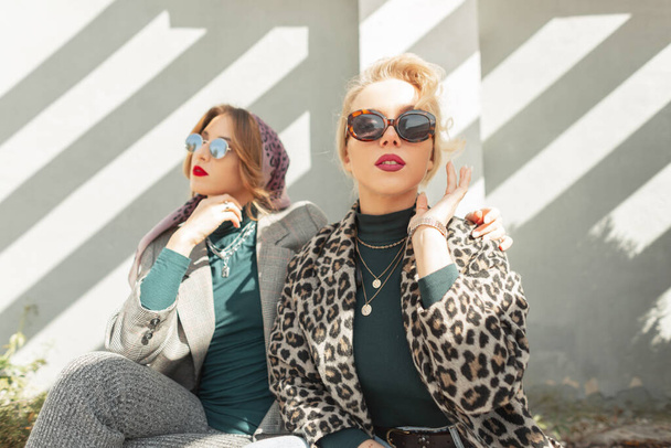 Zwei modische junge Freundinnen mit trendigen Vintage-Sonnenbrillen im modischen Outfit sitzen und posieren im Sonnenlicht auf der Straße neben Betonmauern. Weibliche Kleidung im urbanen Stil - Foto, Bild