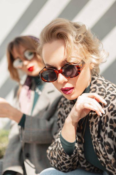 Glamorous young stylish pretty blond woman with vintage sunglasses in fashionable clothes with leopard coat sits with her fashionable girlfriend on the street in the sunlight - Zdjęcie, obraz