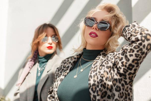 Fashion pretty blonde woman with vintage sunglasses in stylish leopard coat and sweater walks on the street with girlfriend in sunlight. Two trendy urban girls in the city - Photo, Image