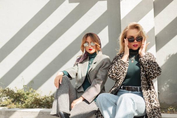Δύο κομψότητα μόδας όμορφα κορίτσια με κομψά ρούχα κάθεται στο δρόμο κοντά σε ένα γκρίζο τοίχο. Αστικό γυναικείο στυλ στολή - Φωτογραφία, εικόνα