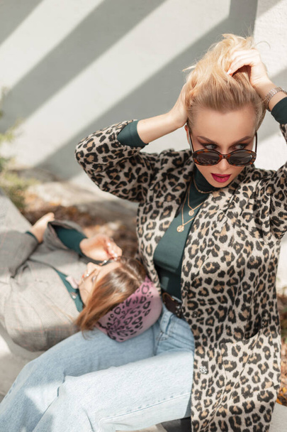 Fashion blonde woman with sunglasses in trendy leopard coat, blue jeans and sweater sits with girlfriend on the street on sunny day. Female casual stylish clothes and beauty - Foto, immagini