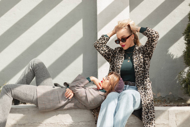 Fashion young pretty girls in stylish autumn outfit with sunglasses, leopard coat, jeans and trendy gray suit sits and rest on the street in sunlight, fashionable female clothes look - Foto, Imagen