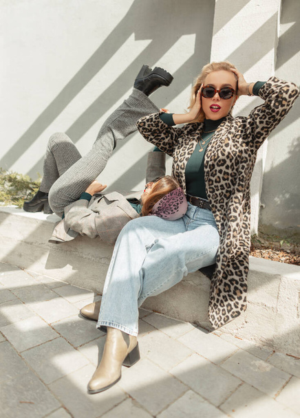 Due divertenti ragazze alla moda hipster con occhiali da sole in un cappotto di leopardo alla moda con jeans e scarpe sono seduti per strada alla luce del sole. Estate fine settimana soleggiato e moda - Foto, immagini