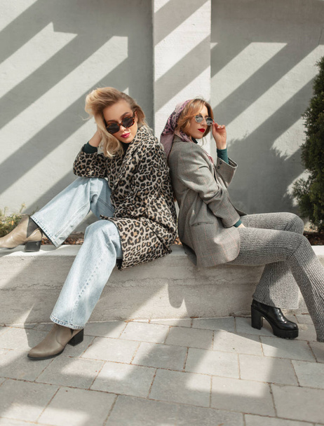 Two fashion girls in trendy spring outfit with sunglasses, leopard coat, jeans, elegant suit and shoes sits in the city - Foto, afbeelding