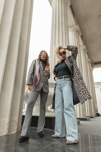 Two fashion young women with trendy autumn vintage clothes stands near a columns of building on the street - Foto, Imagen