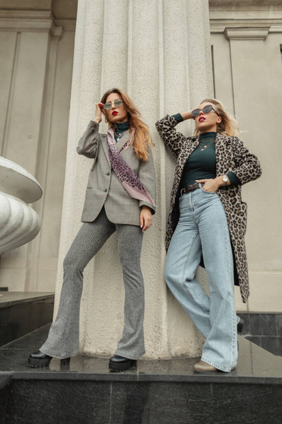 Две элегантные модные девушки в солнечных очках в модной осенней одежде в костюме, пальто, синих джинсах и обуви стоят и позируют возле старинной колонны в городе - Фото, изображение