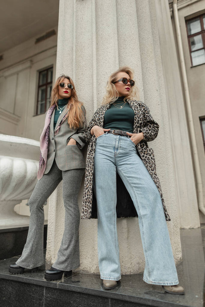 Mooie jonge modieuze meisjes in stijlvolle vintage kleding met jassen, hoge taille bell-bottom jeans en pakken staan vlakbij retro kolommen in de stad - Foto, afbeelding