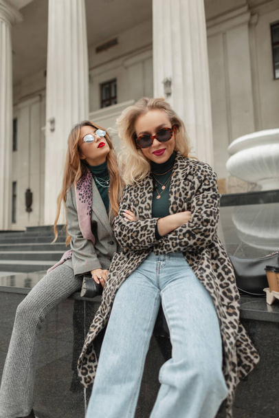 Happy beautiful fashion women in stylish branded autumn clothes with sunglasses, coats and jeans are sitting in the city near the vintage building with columns - Foto, Imagen