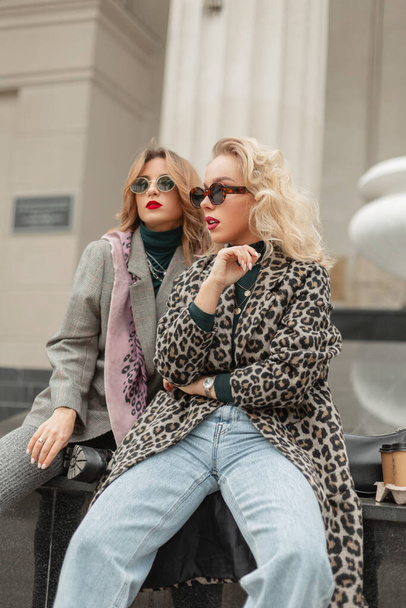 Две модные европейские девушки-хипстерки в очках в модном леопардовом пальто, джинсах и костюме сидят и позируют в городе - Фото, изображение