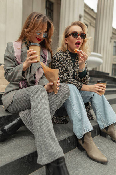 Δύο αστείες νεαρές όμορφες φιλενάδες στη μόδα μοντέρνα ρούχα κάθονται σε σκαλοπάτια, πίνουν καφέ και τρώνε προφιτερόλ στην πόλη. Ευτυχισμένος τρόπος ζωής και ταξίδια - Φωτογραφία, εικόνα