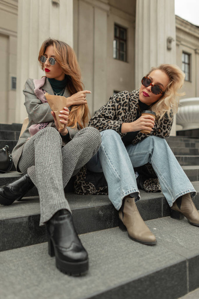 Two glamorous young hipster girls drinking coffee and sitting on the steps in the city - Foto, Imagen
