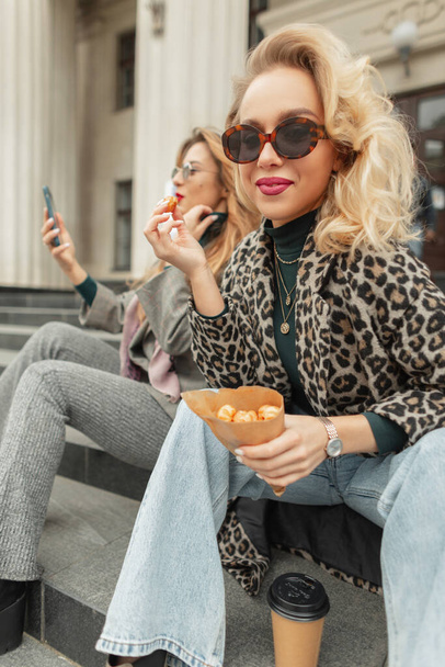 Feliz moda joven hermosa mujer con gafas de sol en ropa de moda con estilo con abrigo de leopardo y vaqueros comiendo un profiteroles y bebe un café en la ciudad. Vida urbana de felicidad - Foto, Imagen