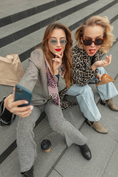Αστεία ευτυχία όμορφη νεαρά καυκάσια κορίτσια με γυαλιά ηλίου σε μοντέρνα ρούχα κάθονται στα σκαλιά, πίνουν καφέ, τρώνε profiteroles και να πάρετε μια φωτογραφία selfie στο τηλέφωνο, top view - Φωτογραφία, εικόνα