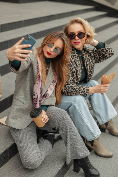 Δροσερά νεαρά hipster κορίτσια με μοντέρνα ρούχα κάθονται στα σκαλιά πίνοντας καφέ και λαμβάνοντας selfies στην πόλη - Φωτογραφία, εικόνα