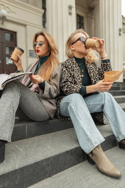 Стильні красиві гламурні дівчата в модному одязі, що сидять на сходах, п'ють каву в місті і читають журнал
 - Фото, зображення