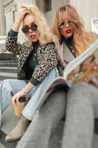 Mujeres hermosas de moda en ropa retro con estilo se sientan en la ciudad en los escalones, beber café y leer una revista - Foto, imagen