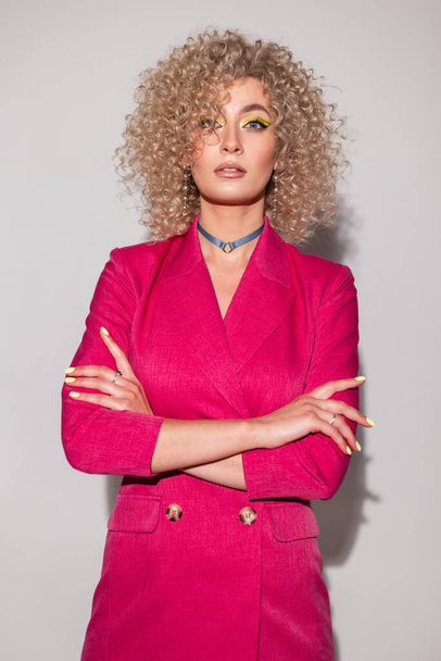 Beautiful glamorous business woman with a curly afro hairstyle in a pink suit stands in the studio and looks into the camera. Girl with colorful bright makeup - 写真・画像