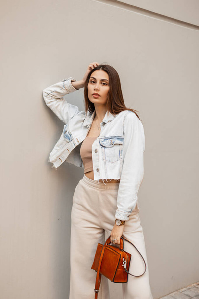 Trendy young beautiful woman with fashion blue denim jacket, top, pants and handbag stands and poses near a gray wall - Foto, Bild