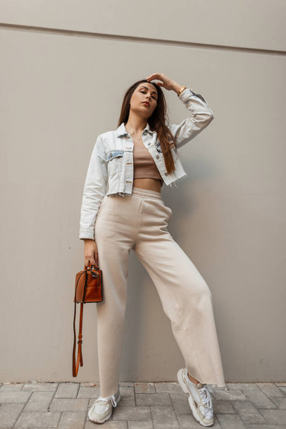 Stylová krásná mladá dívka v módním oblečení s džínovou bundou, kabelkou, širokými kalhotami a tenisky stojící u šedé zdi ve městě - Fotografie, Obrázek