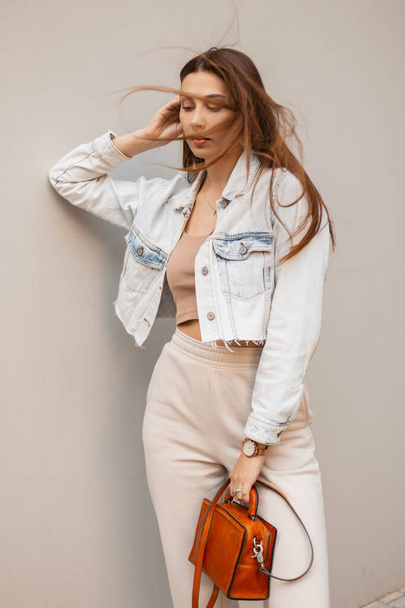Красива молода модна жінка з брюнеткою в джинсовій куртці зі шкіряною сумкою стоїть біля сірої стіни в місті
 - Фото, зображення