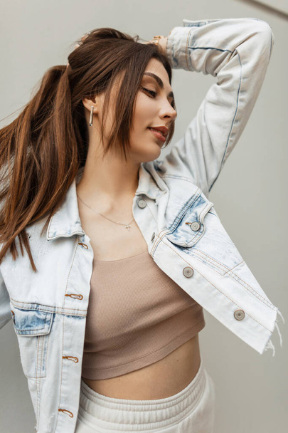 Urban portrait of young pretty brunette woman with hairstyle in fashion blue jeans jacket and top poses on the street near a gray wall - Fotoğraf, Görsel