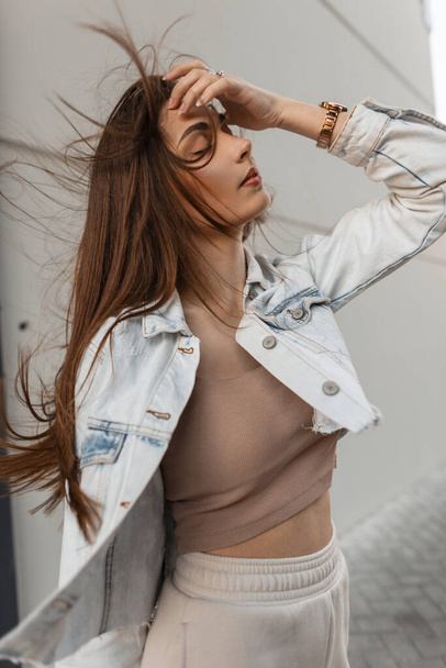 Retrato urbano de la joven belleza de moda con pelo moreno con chaqueta jeans camina por la calle cerca de una pared gris - Foto, Imagen