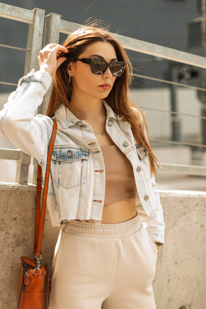 Όμορφη νεαρή μοντέρνα κορίτσι μοντέλο με μοντέρνα μαύρα γυαλιά ηλίου σε μοντέρνα ρούχα denim με μια τσάντα στέκεται στην πόλη - Φωτογραφία, εικόνα