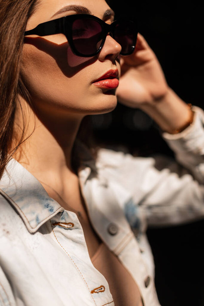 Retrato glamouroso na moda de uma bela jovem com óculos de sol elegantes em uma jaqueta de jeans casual na rua à luz do sol - Foto, Imagem