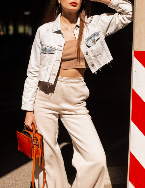 Modna szczupła dziewczyna w stylowych markowych ubraniach casual z dżinsową kurtką, spodniami na dole dzwonka, top i torebką w mieście w świetle słonecznym - Zdjęcie, obraz