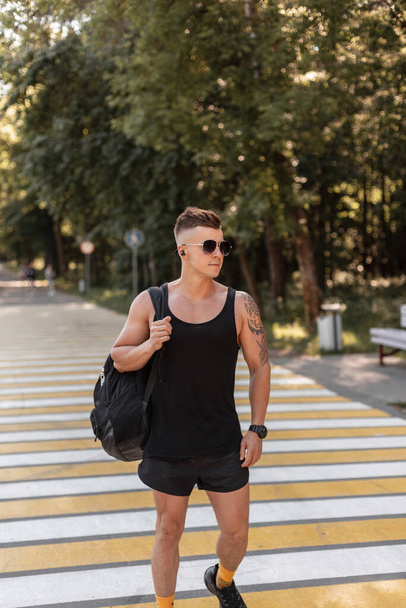 Sporty handsome young man with sunglasses and summer style clothes with shorts, tank top, and sneakers with a backpack walks in the city and goes to practice - Foto, afbeelding