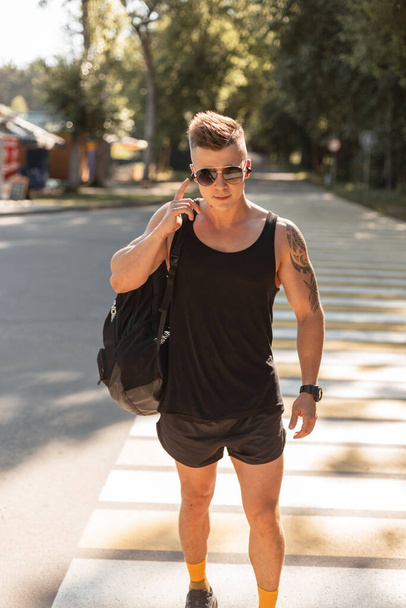 Sports handsome hipster man with hairstyle and sunglasses in fashionable black summer clothes with a backpack walks outdoors and listens to music in wireless headphones - Foto, Imagen