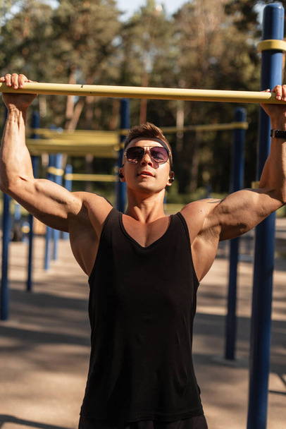 Athletic strong guy with wireless earphones and sunglasses in black sportswear pulls himself up on horizontal bars outdoors. Man exercising, healthy lifestyle - Photo, Image