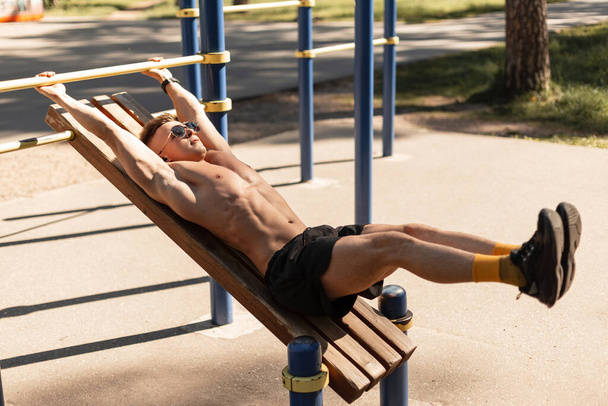 Young handsome man athlete with muscular body doing leg raise clap on a wooden bench outdoors on sunny day - Фото, зображення