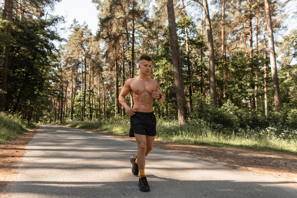 Knappe jonge atleet man met naakt gespierde romp loopt op de weg in het bos. Buiten rennen en een gezonde levensstijl - Foto, afbeelding