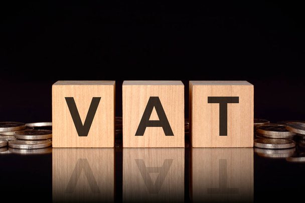 vat - short for Value Added Tax. text on wood cubes with coins, black background, business concept. the inscription on the cubes is reflected from the surface of the black table. front view - Photo, Image