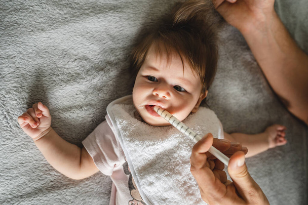 Close up on small caucasian baby four months old and hand of unknown father giving her medicine antibiotics while lying on bed at home parenthood and health issues liquid drugs oral syringe concept - 写真・画像