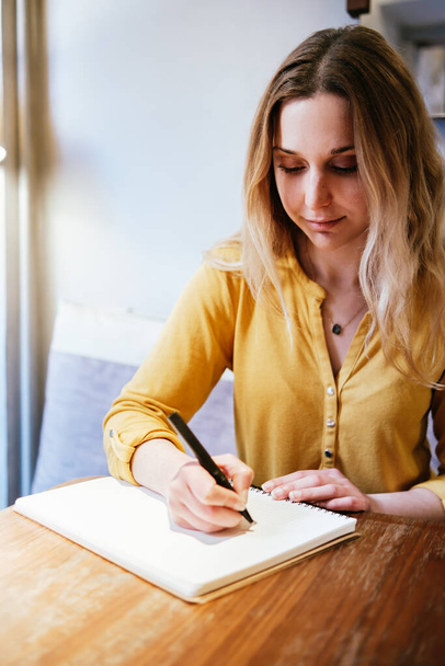 νεαρή λευκή γυναίκα που κάθεται σε καφετέρια και γράφει στο χέρι σε ένα χάρτινο τετράδιο - Φωτογραφία, εικόνα