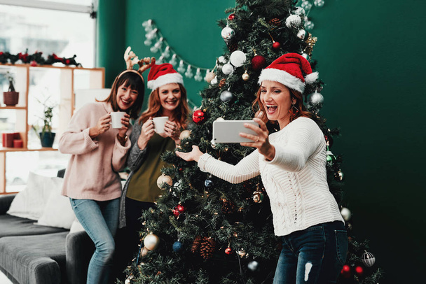 木の近くに立ってください。3人の魅力的な中年女性の切り取られたショットクリスマスの時間の間に自宅で携帯電話と一緒に自分の肖像画を撮る. - 写真・画像
