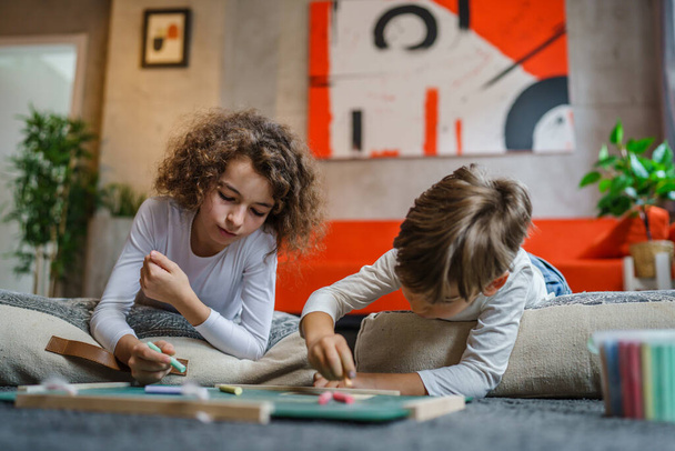 small boy and girl drawing on the green board at home two children brother and sister siblings or friends lying on the floor using chalk leisure and education real people family concept copy space - Valokuva, kuva