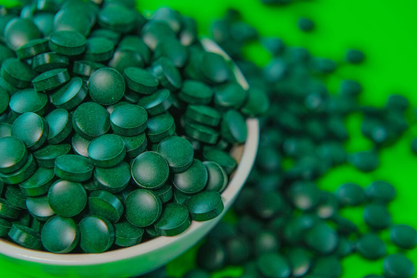 Spirulina pillereitä pyöreässä kulhossa. Vihreät spirulina-tabletit vihreällä taustalla. Spirulina-levät tabletit. Vitamiinit ja ravintolisät. Super ruoka. Ravintolisät terveelliseen elämäntapaan - Valokuva, kuva