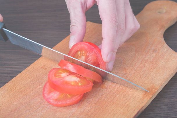 Slice Tomatoes in Half. Red tomato on the chopping Board, in the kitchen. Female Chef cuts one red tomato with a knife on wooden cutting board. The process of cooking, close-up. - Фото, изображение