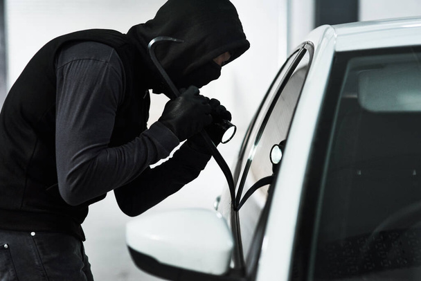 Hes knows no bounds. Shot of a masked man using a steel weapon to break into a car inside a parking lot. - Foto, Imagen