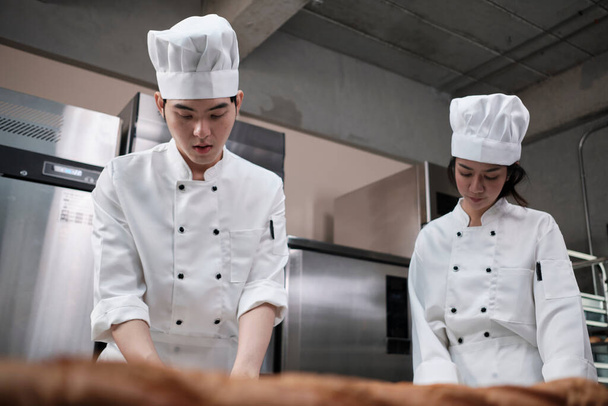 Twee professionele Aziatische koks in witte kok uniformen en schorten kneden deeg en eieren, bereiden brood, koekjes en vers bakkerijvoedsel, bakken in een oven in een restaurant roestvrij stalen keuken. - Foto, afbeelding