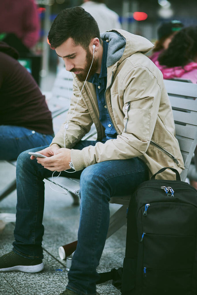 時間を渡すために技術を使用しています。バスの駅に座っている間、彼の携帯電話で音楽を聞いてハンサムな若い男の完全な長さのショット. - 写真・画像