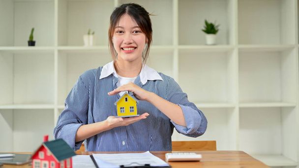 Attractive Asian young female real estate agent or realtor showing a tiny house model to camera, sitting at office desk. Property investment, House Insurance. - Photo, Image